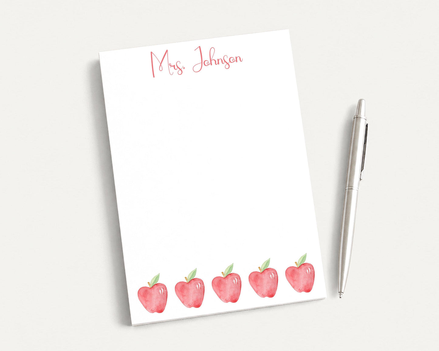 a white notepad with a watercolor illustration of five red apples, accompanied by a pen on a white surface.