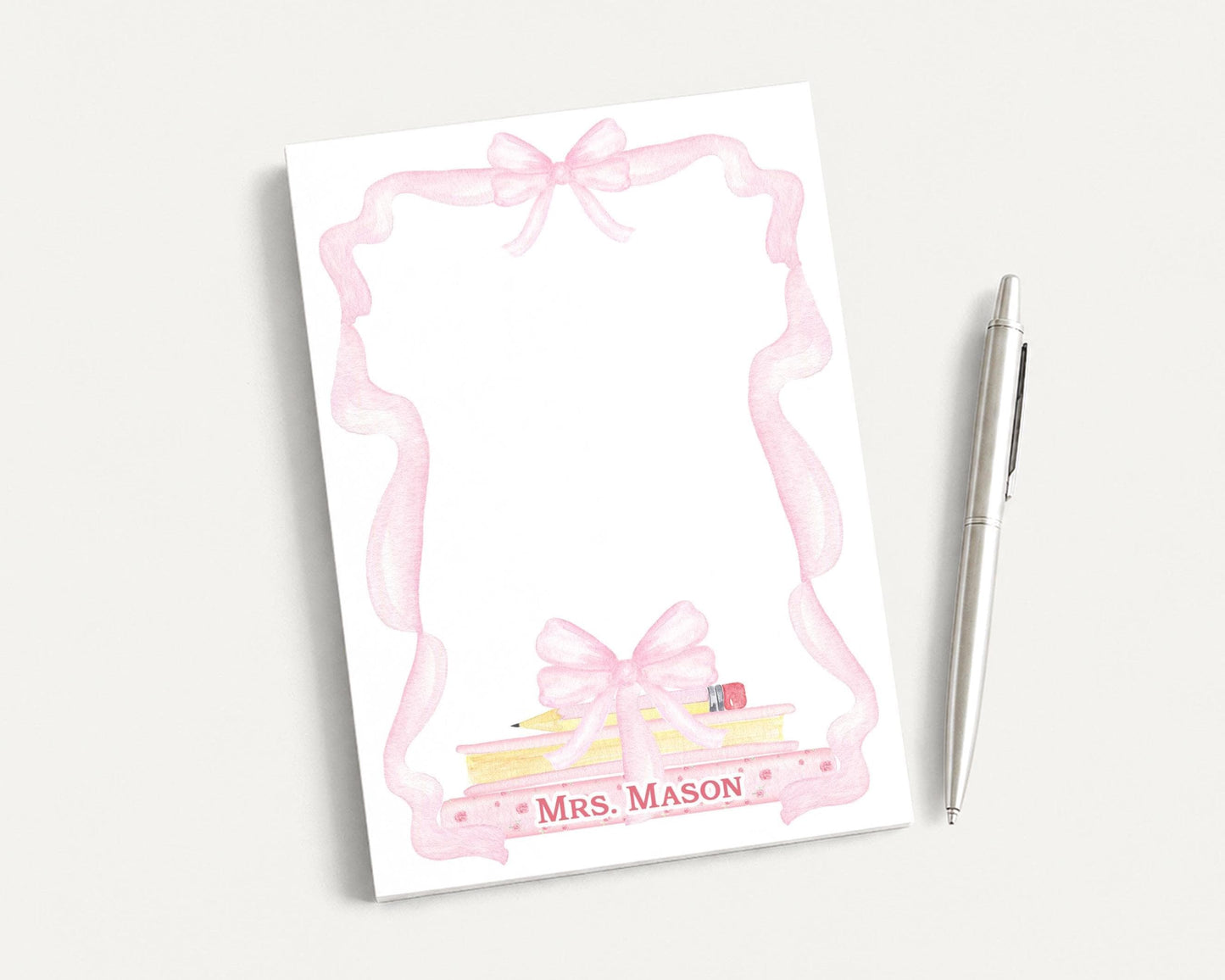A white notepad with a pink ribbon frame and a pencil on top, along with a pen, is placed on a white surface.