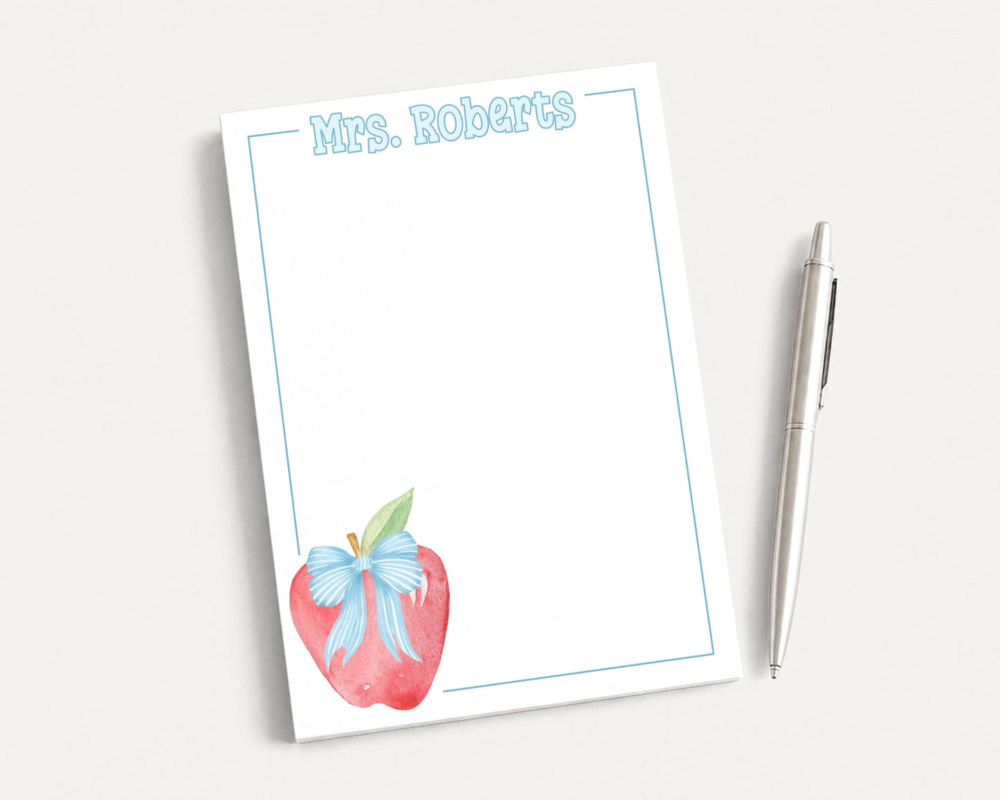 a blank notepad with a watercolor illustration of an apple on the cover, accompanied by a pen on a white surface.