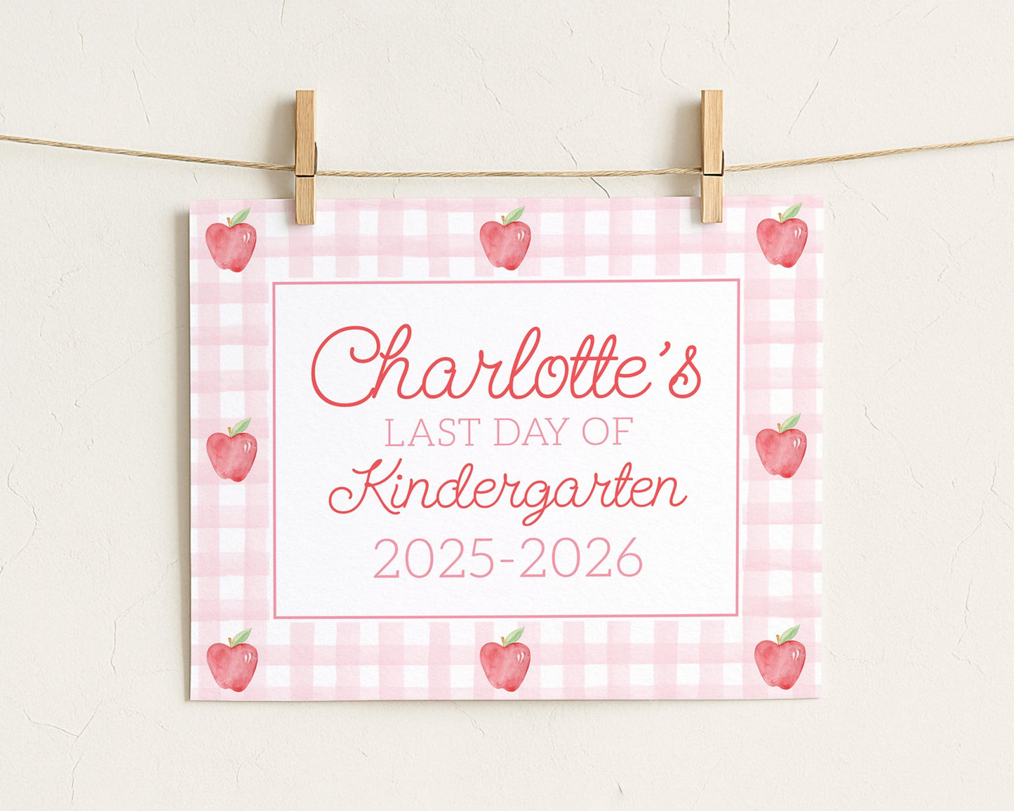A pink and white checkered sign with the text "Charlotte's LAST DAY OF Kindergarten 2015-2026" and several red apples on a string.