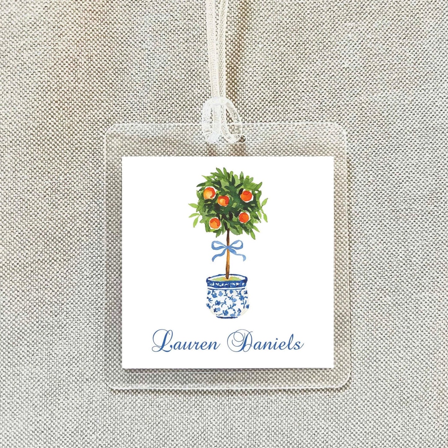 A square tag with a watercolor illustration of a potted tree with oranges on top, hanging from a string.