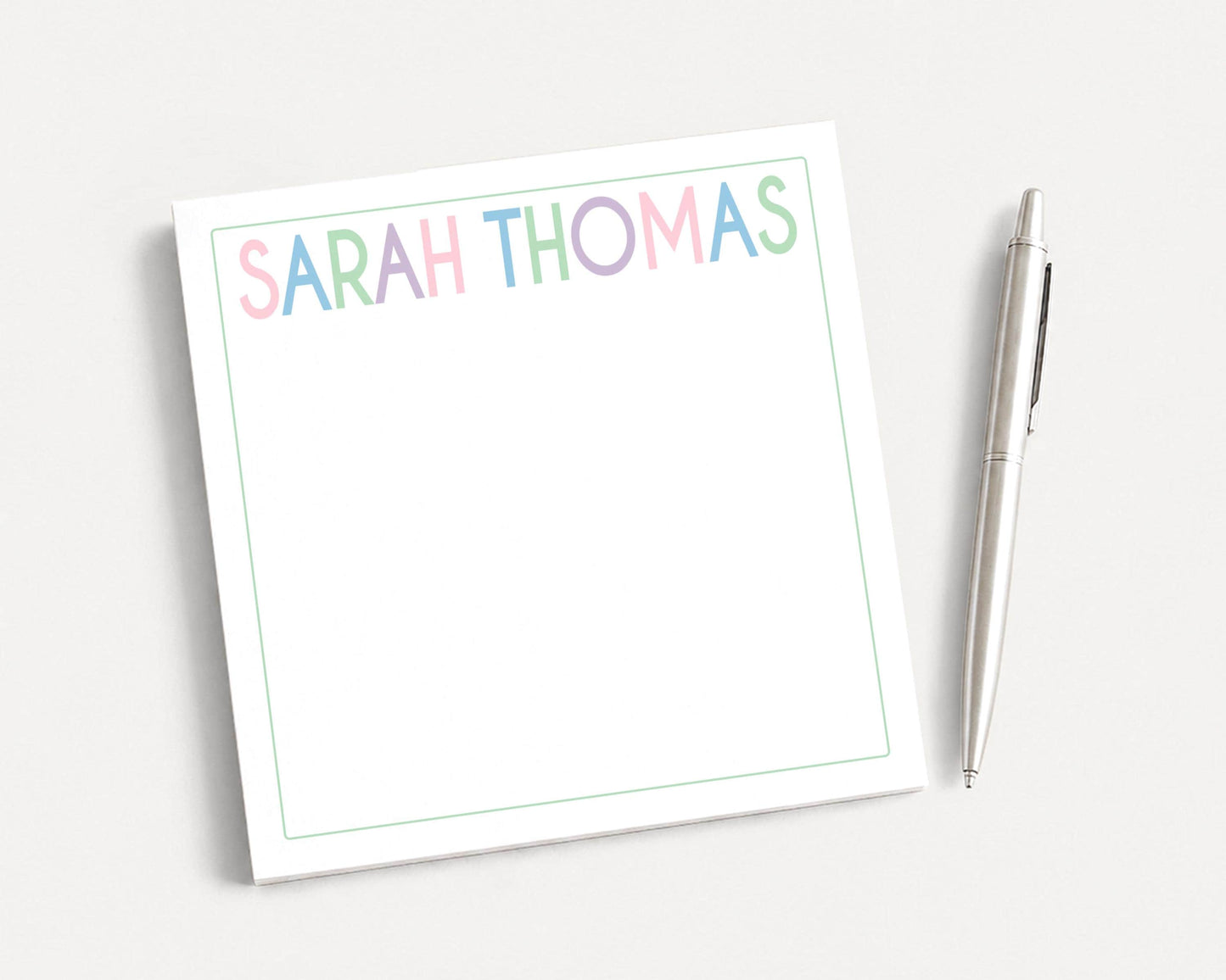 A white notepad with the name "SARAH THOMAS" written on it, along with a pen, is placed on a plain white surface.