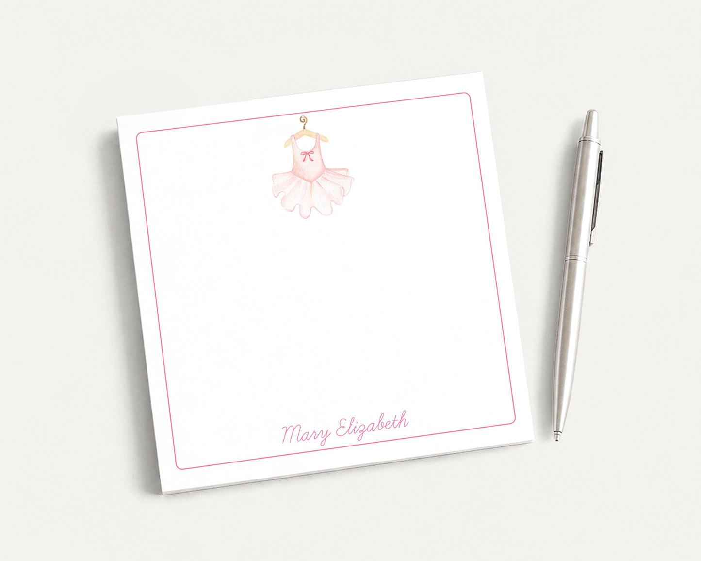 A white notepad with a pink ballet tutu illustration and the name "Mary Elizabeth" written on it, along with a pen on a plain white background.