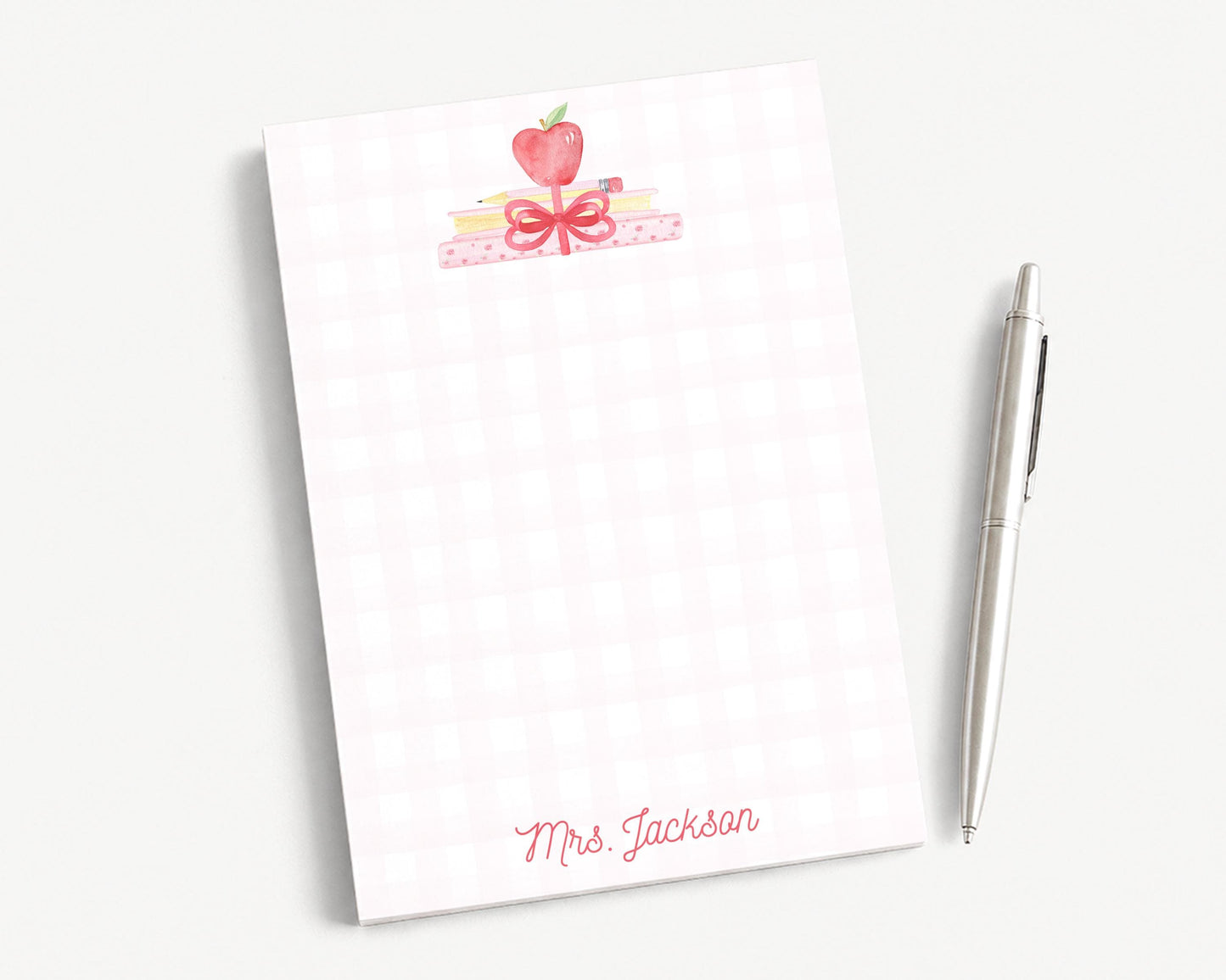 A white notepad with a pink and red design, featuring a heart and a bow, along with a pen and a pencil on a white surface.