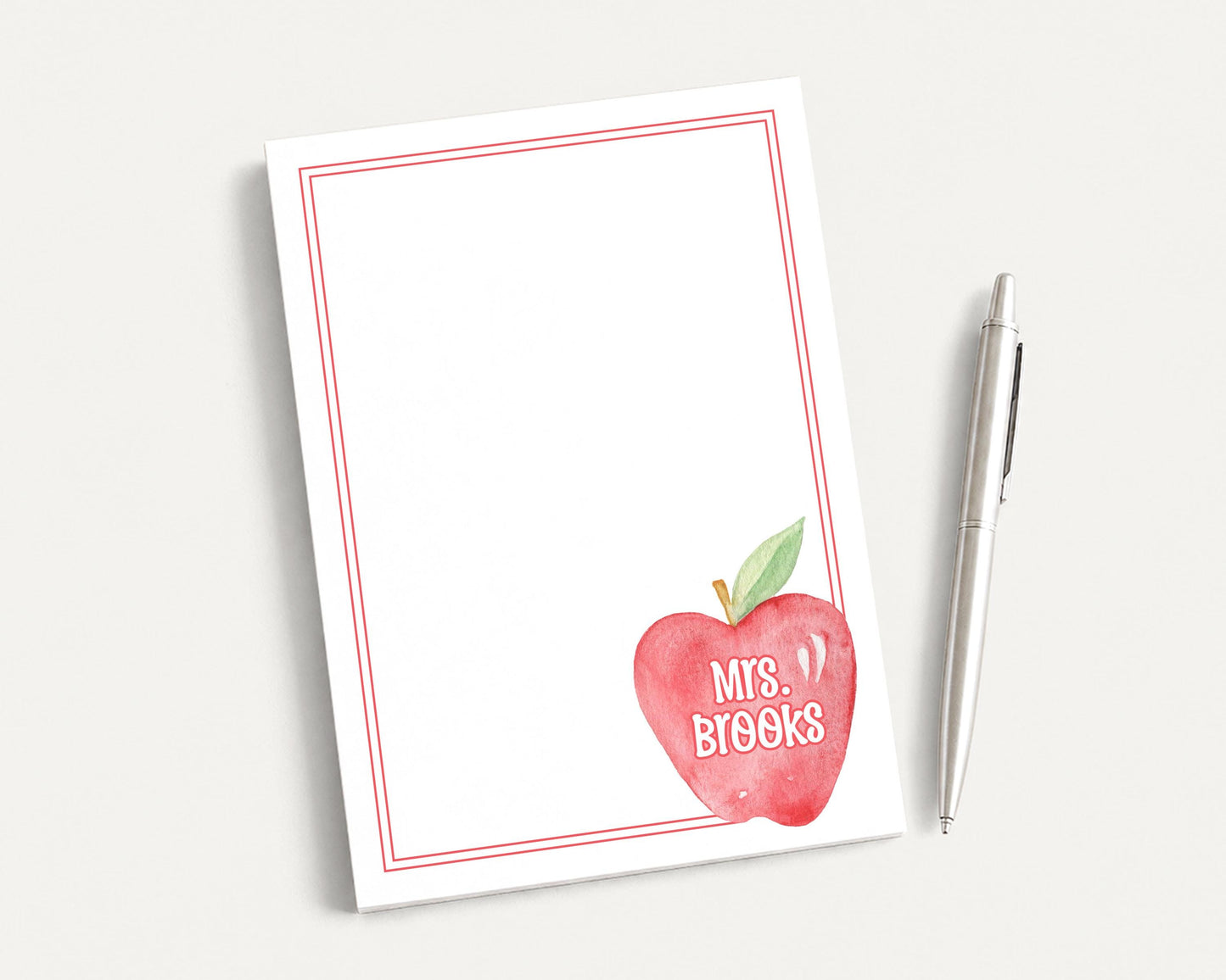 A white notepad with a red border and an apple graphic, along with a pen, is placed on a white surface.