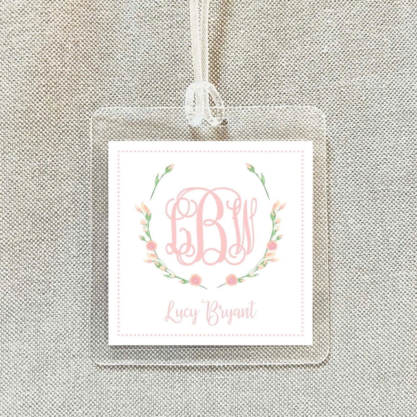 A clear square tag with a monogrammed floral design and the name "Lucy Bryant" printed on it.