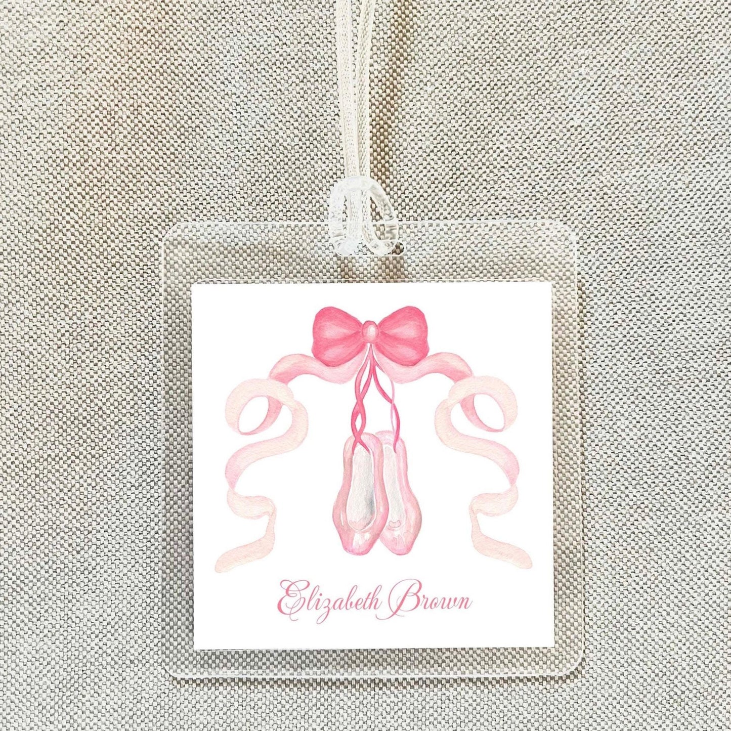 A clear name tag with a pink ribbon and ballet slippers printed on it, hanging on a fabric background.