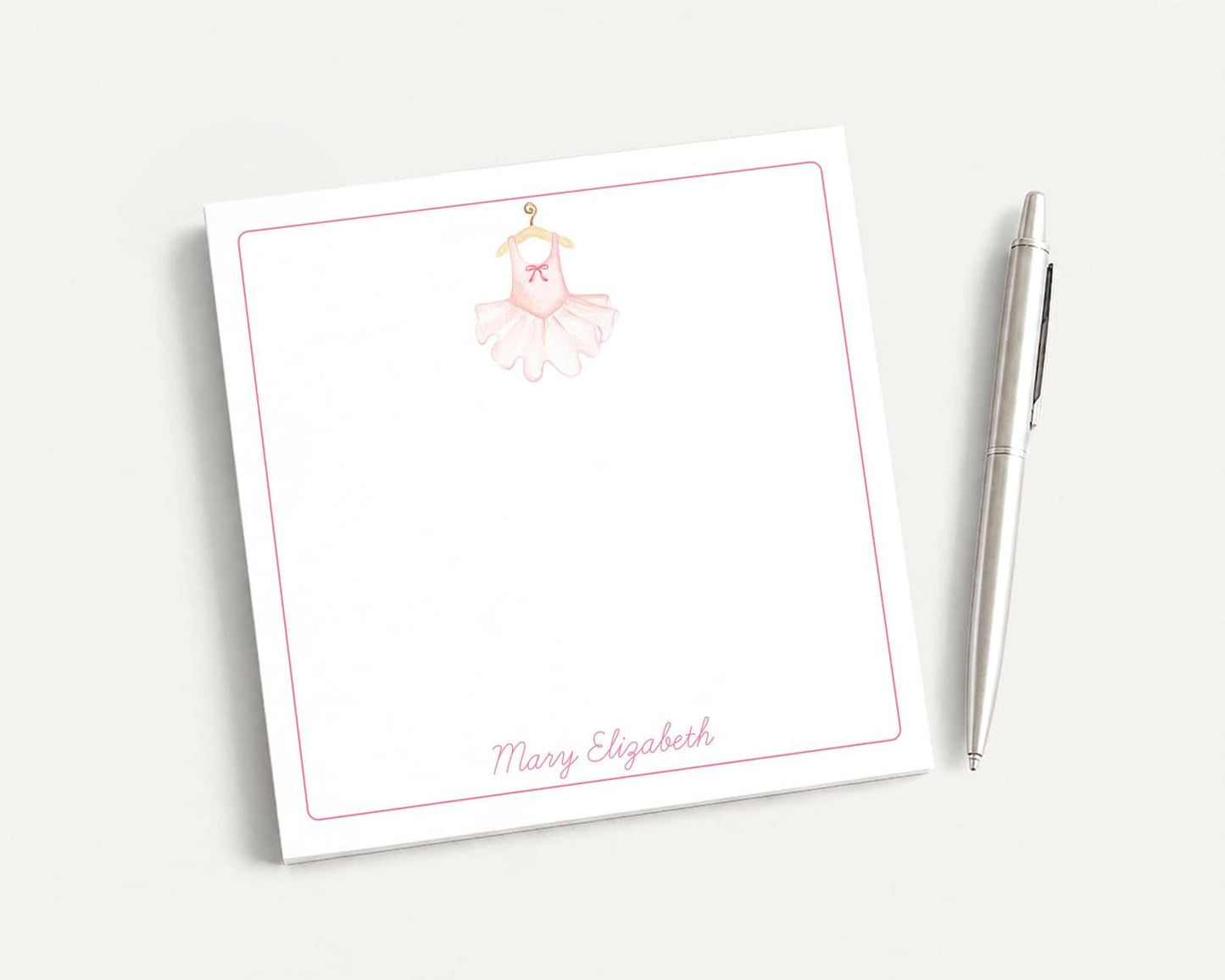 A white notepad with a pink ballet tutu illustration and the name "Mary Elizabeth" written on it, along with a pen on a white surface.