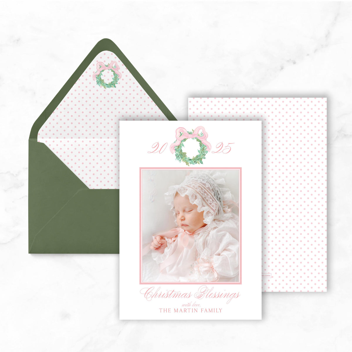 A Christmas card featuring a baby in a white dress and a wreath, with a green envelope and a white card with pink polka dots on a marble background.