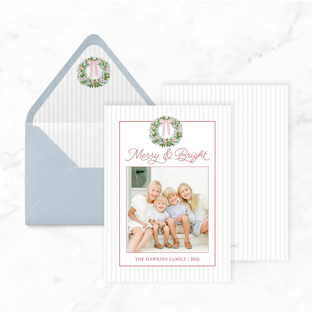 A white card with a photo of a family sitting on a porch, and a blue envelope with a wreath design on it, both placed on a marble surface.