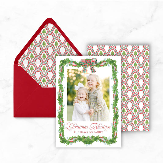A Christmas card featuring two young girls in a wreath frame, with a red envelope and a patterned card visible in the background.