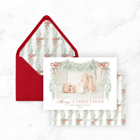 A Christmas card featuring a baby photo with a red envelope decorated with a pattern of green and red bows.
