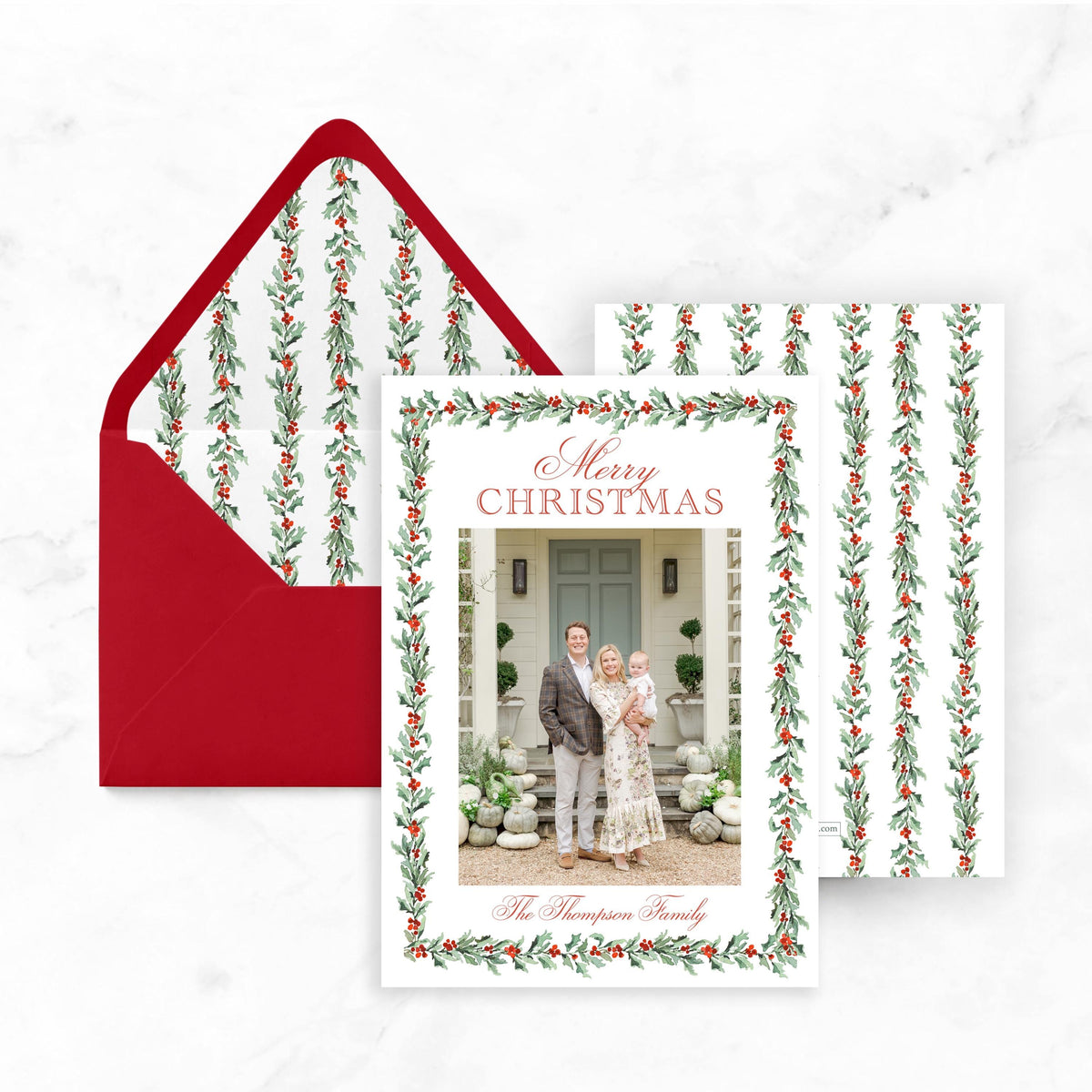 A Christmas card with a photo of a family, including a man, a woman, and a child, is displayed next to an envelope with a red flap and a white background.