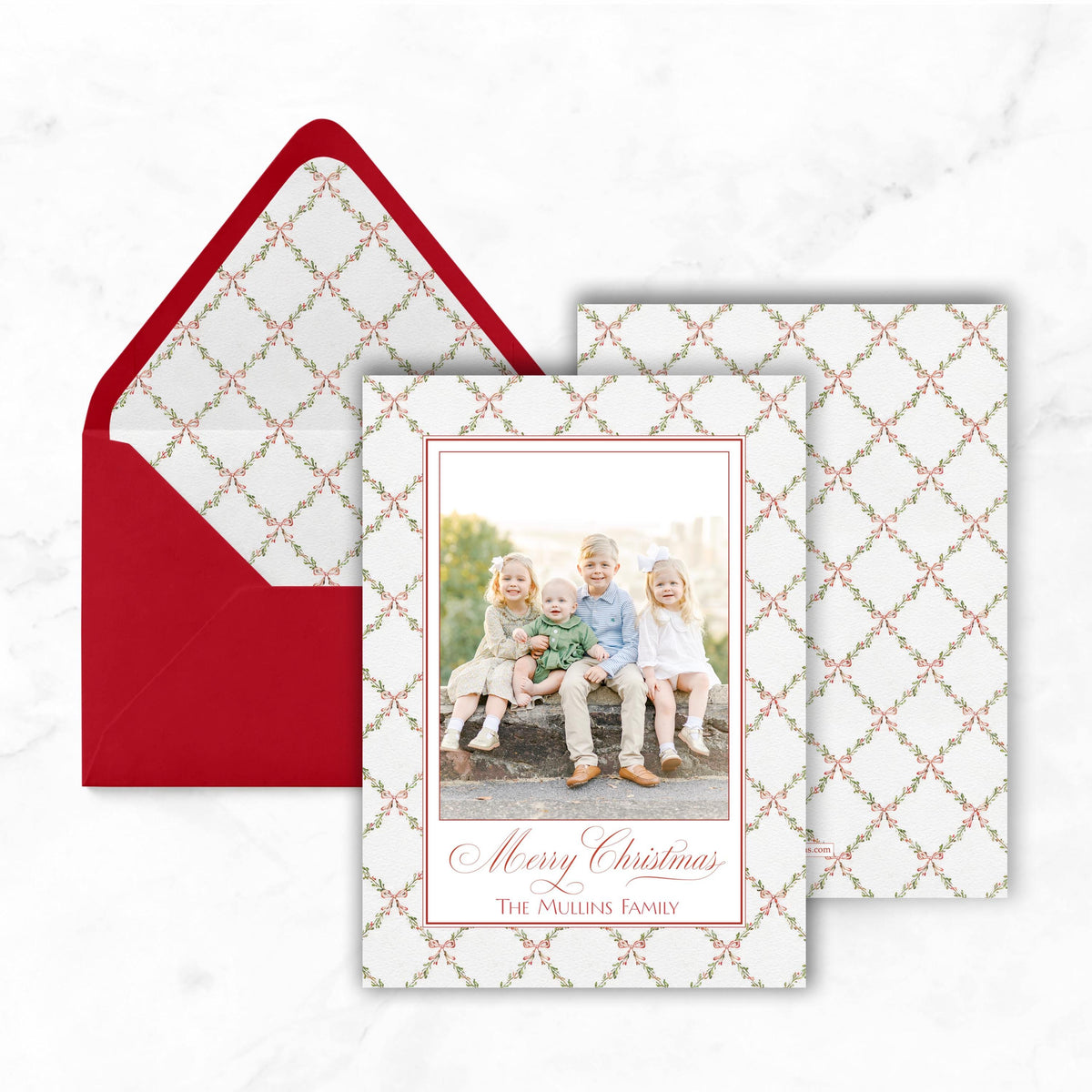 A Christmas card with a photo of four children sitting on a bench, placed inside a red envelope against a white marble background.