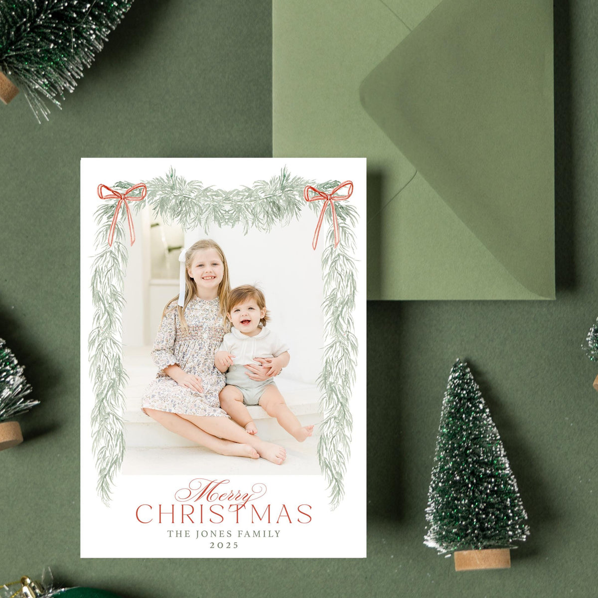A Christmas card with a photo of two children, surrounded by small green Christmas trees, ornaments, and a green envelope on a dark green background.