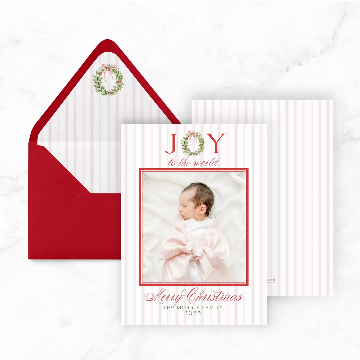 A Christmas card with a photo of a baby and the text "Joy to the world" and "Merry Christmas" printed on it, placed next to an open red envelope with a wreath decoration.
