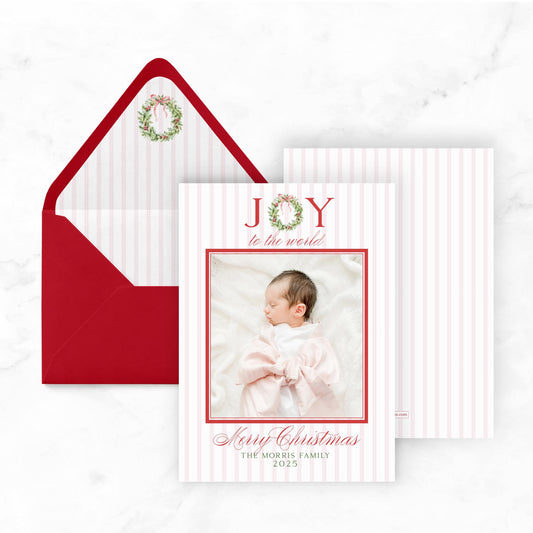 A Christmas card with a photo of a baby and the text "Joy to the world" and "Merry Christmas" printed on it, placed next to an open red envelope with a wreath decoration.