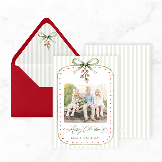 A Christmas card with a photo of three children sitting on a bench, and a bow with a mistletoe and berries on top. The card is placed next to an open red envelope with a bow.