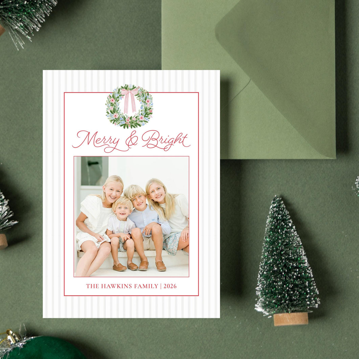 a green envelope, a white card with a photo of three children, and several small green Christmas trees and ornaments arranged on a green background.