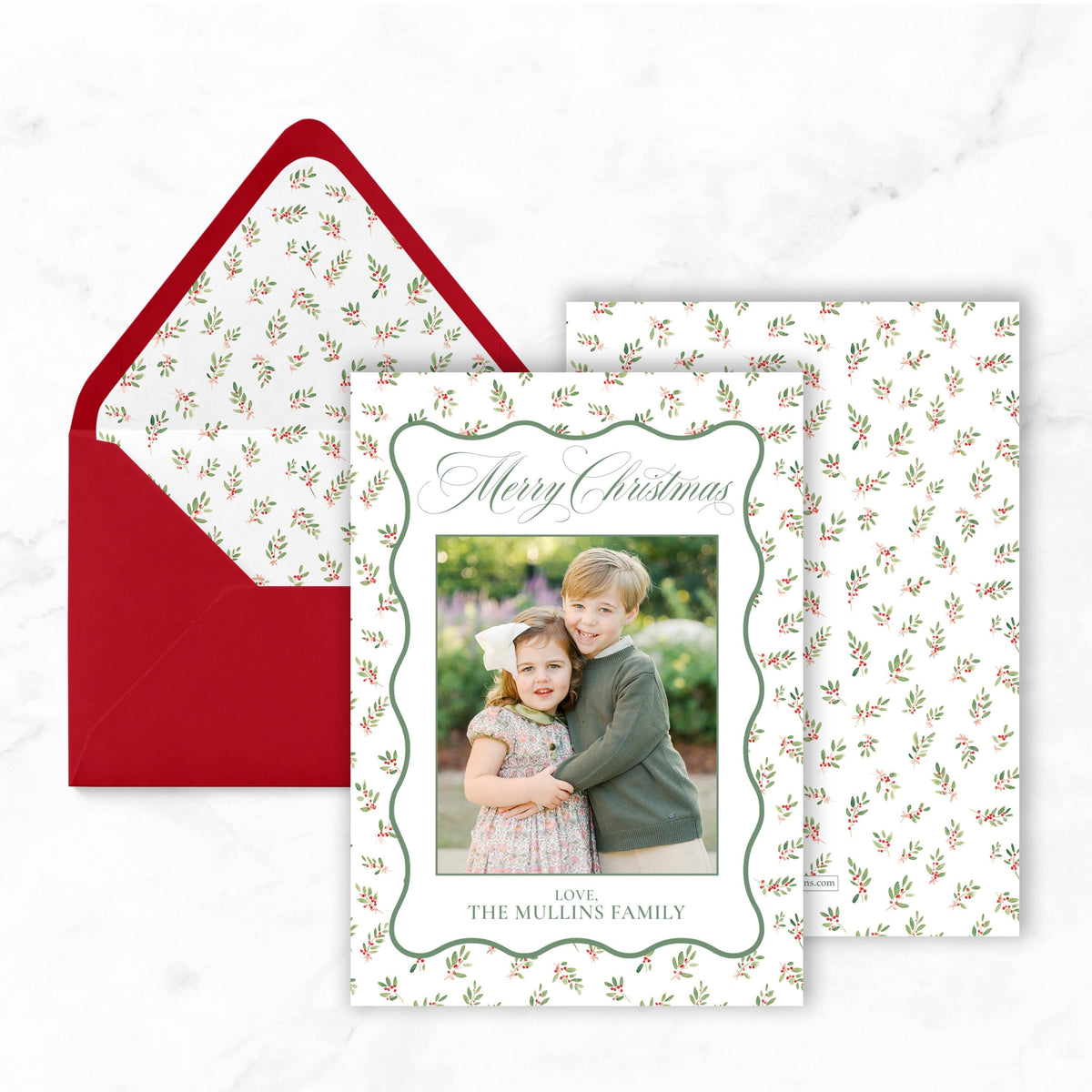 A Christmas card with a photo of two children, one boy and one girl, is displayed in a red envelope with a white floral pattern.