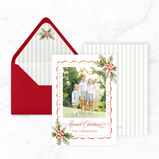 A Christmas card with a photo of three children standing in a field, surrounded by holly leaves and berries, and a red envelope with a white and red striped pattern.