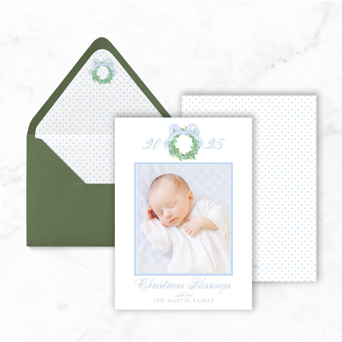 A white card with a photo of a sleeping baby, a green envelope, and a blue bow on a white marble background.