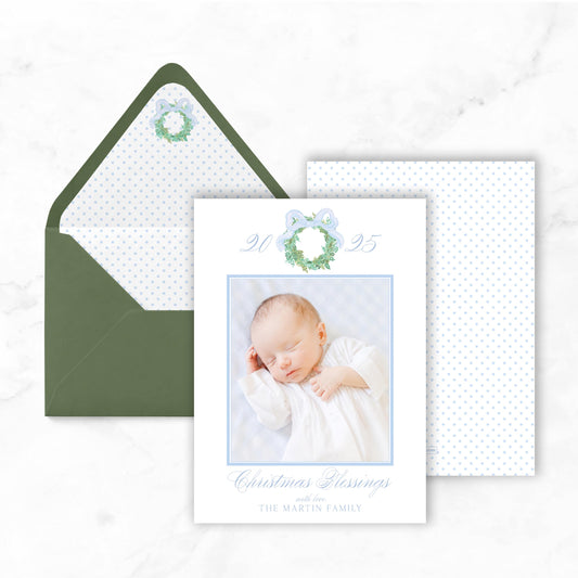 A white card with a photo of a sleeping baby, a green envelope, and a blue bow on a white marble background.
