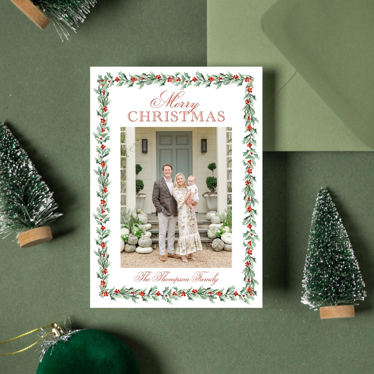 a Christmas card with a photo of a family, surrounded by Christmas decorations such as small trees, ornaments, and a green envelope.