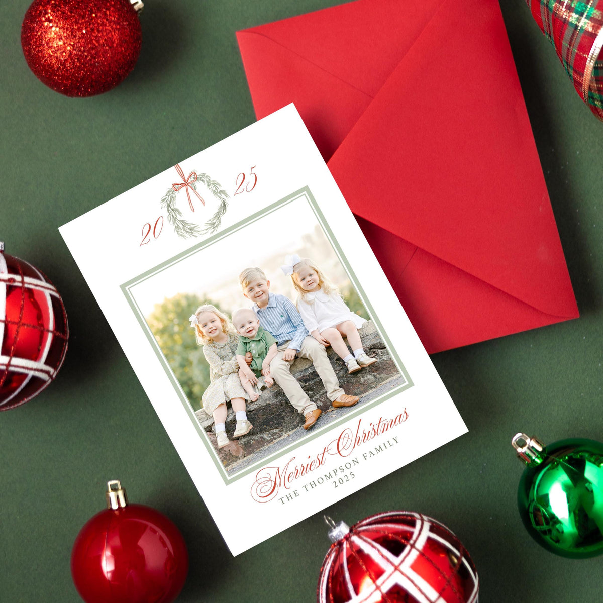 a Christmas card with a photo of three children, surrounded by ornaments and a red envelope on a green background.