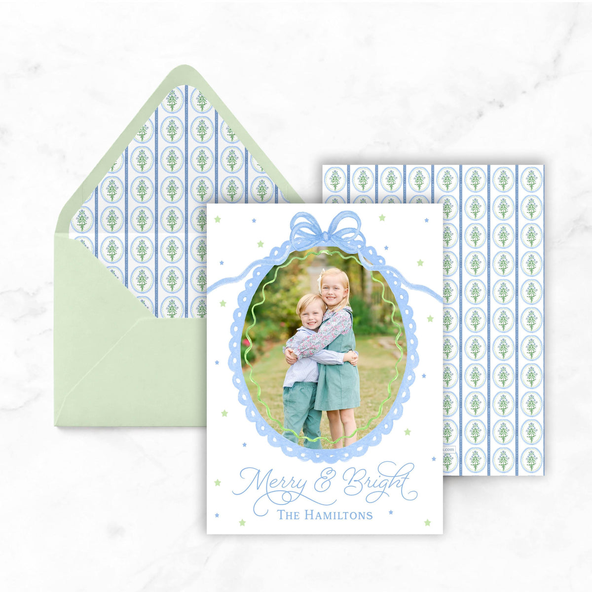 A holiday card featuring two young girls embracing in a garden setting, with a bow around their necks. The card has a photo of the girls and the text "Merry & Bright The Hamiltons".