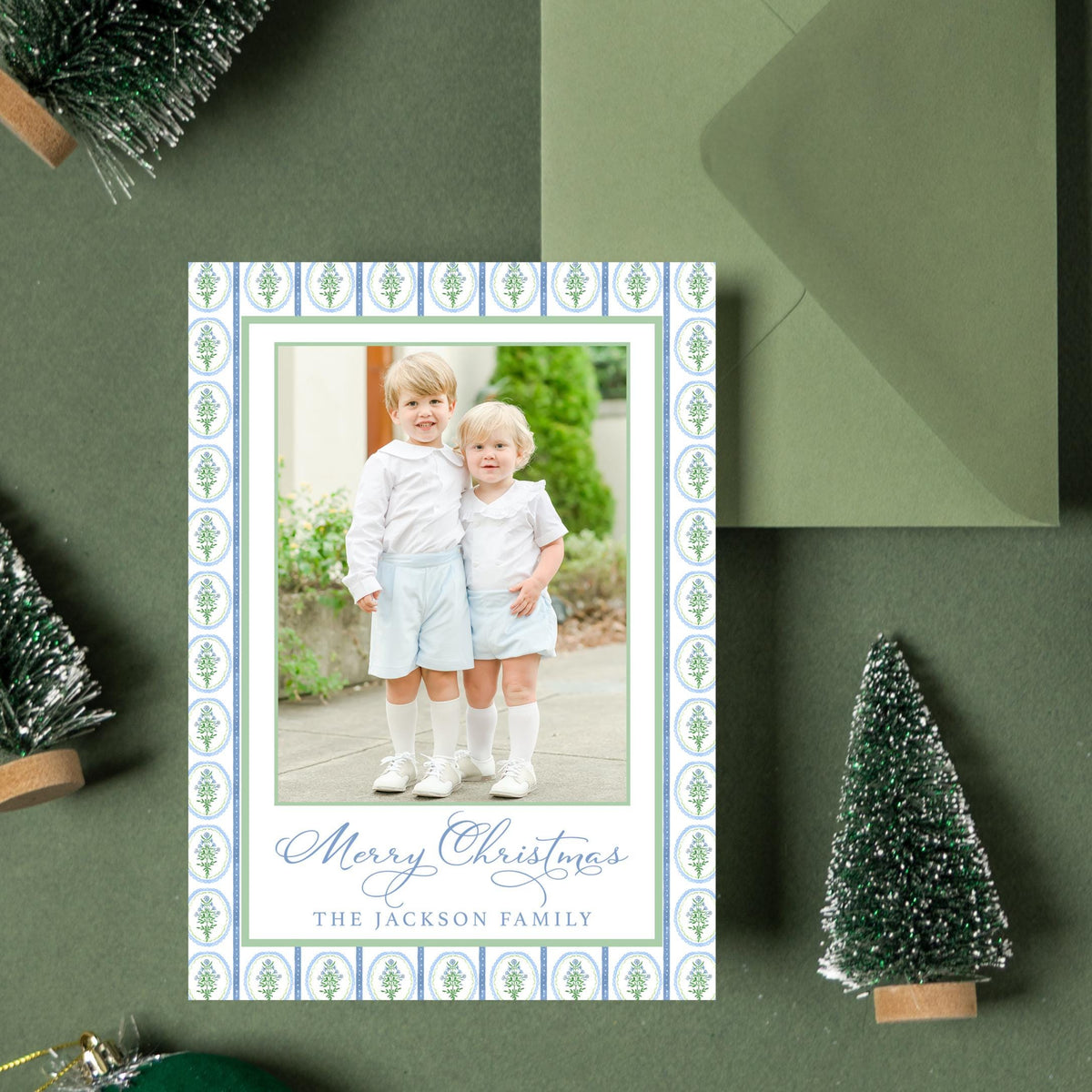 A Christmas card with a photo of two children is surrounded by small green Christmas trees and ornaments on a dark green background.