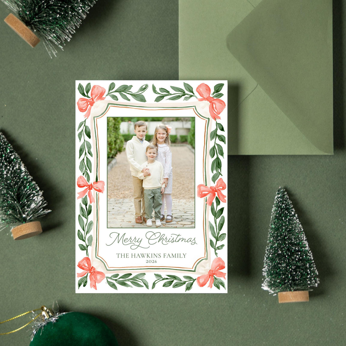 a Christmas card with a photo of a family, surrounded by green Christmas trees, ornaments, and a green envelope.