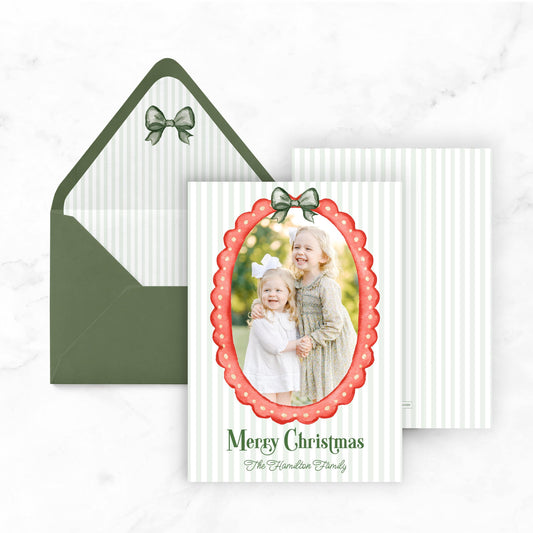 A Christmas card with a photo of two young girls, one wearing a white dress and the other in a pink dress, framed by a red border and a green bow. The card is placed next to an open green envelope with a bow on top.