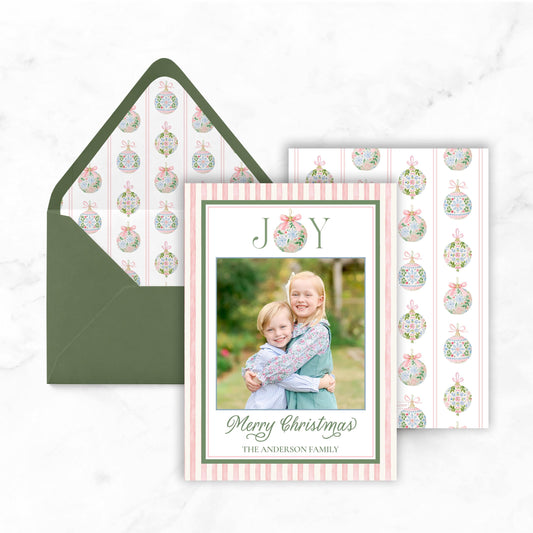 a Christmas card with a photo of two children, one girl and one boy, embracing each other. The card is placed on top of an envelope with a pattern of Christmas ornaments.