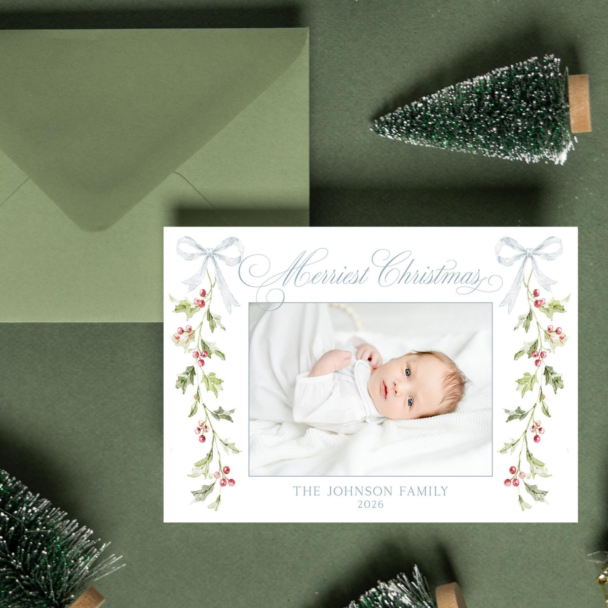 A Christmas card with a photo of a baby is surrounded by green Christmas trees, ornaments, and a green envelope on a green background.
