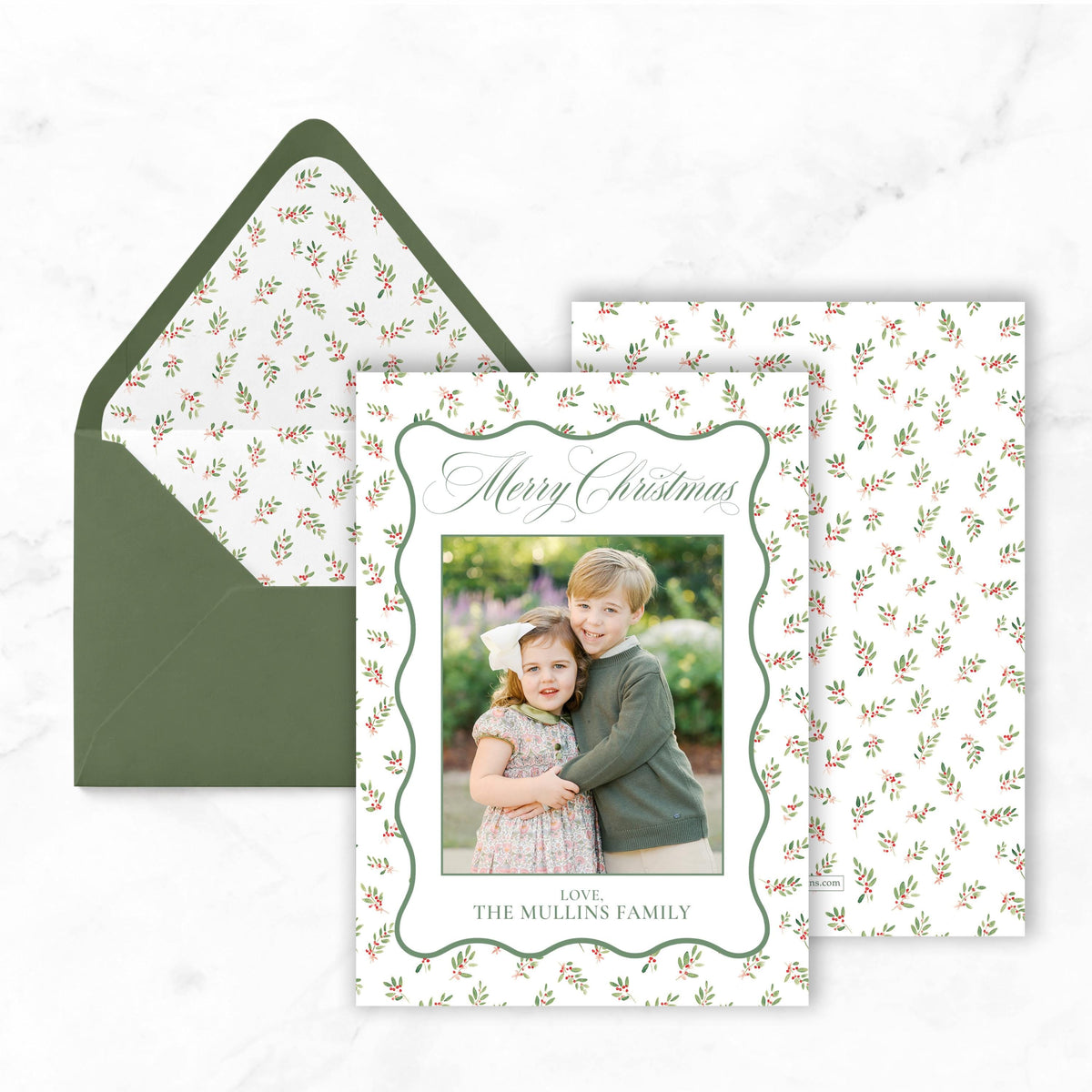 A Christmas card with a photo of two children, likely siblings, and the text "Merry Christmas" and "Love, The Mullins Family" printed on it. The card is placed on top of an envelope with a similar pattern.