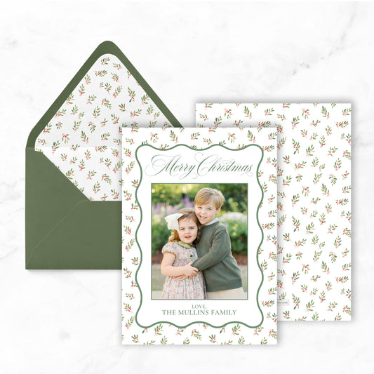 A Christmas card with a photo of two children, likely siblings, and the text "Merry Christmas" and "Love, The Mullins Family" printed on it. The card is placed on top of an envelope with a similar pattern.