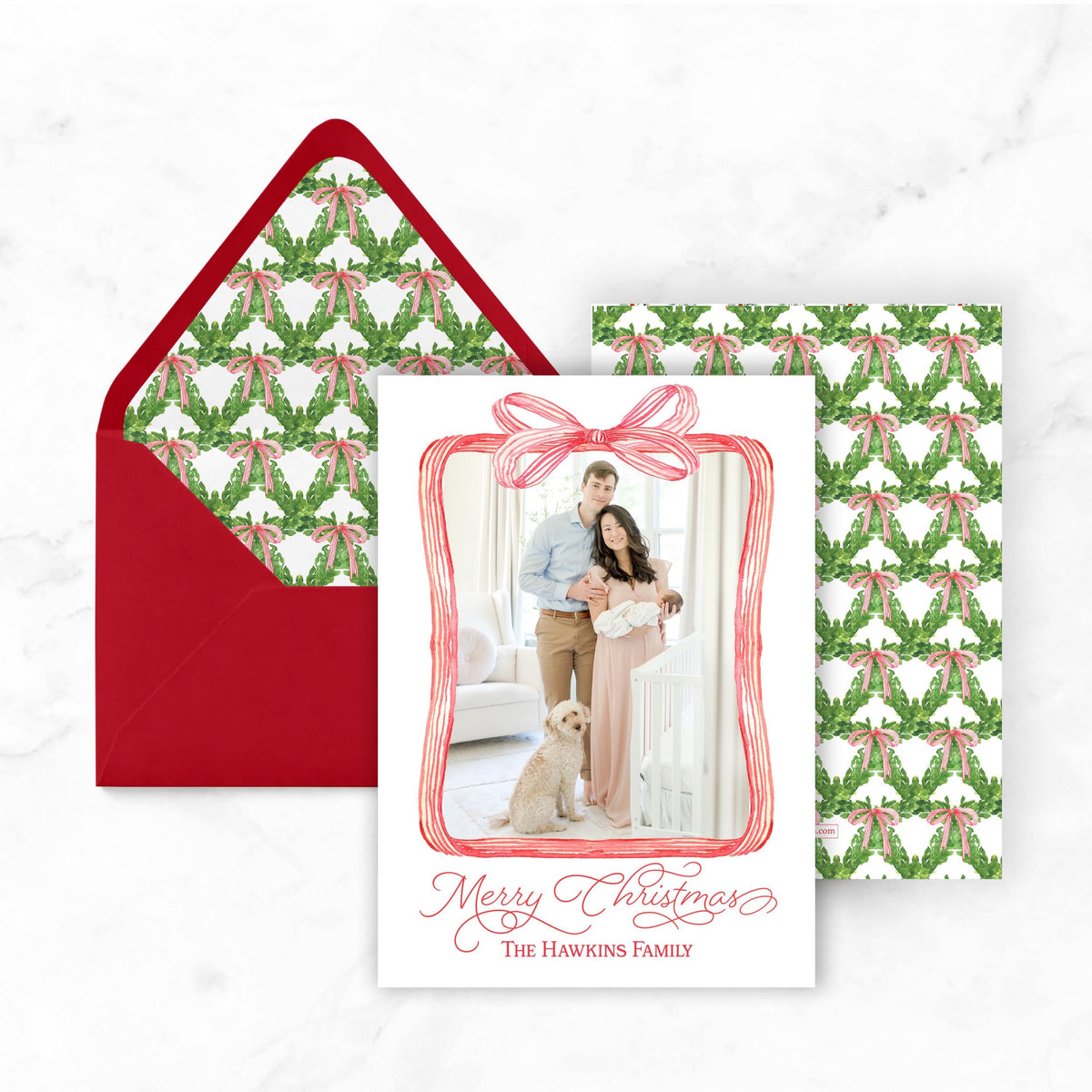 A Christmas card featuring a family photo with a bow, placed on top of a red envelope with a pattern of green leaves and red bows.