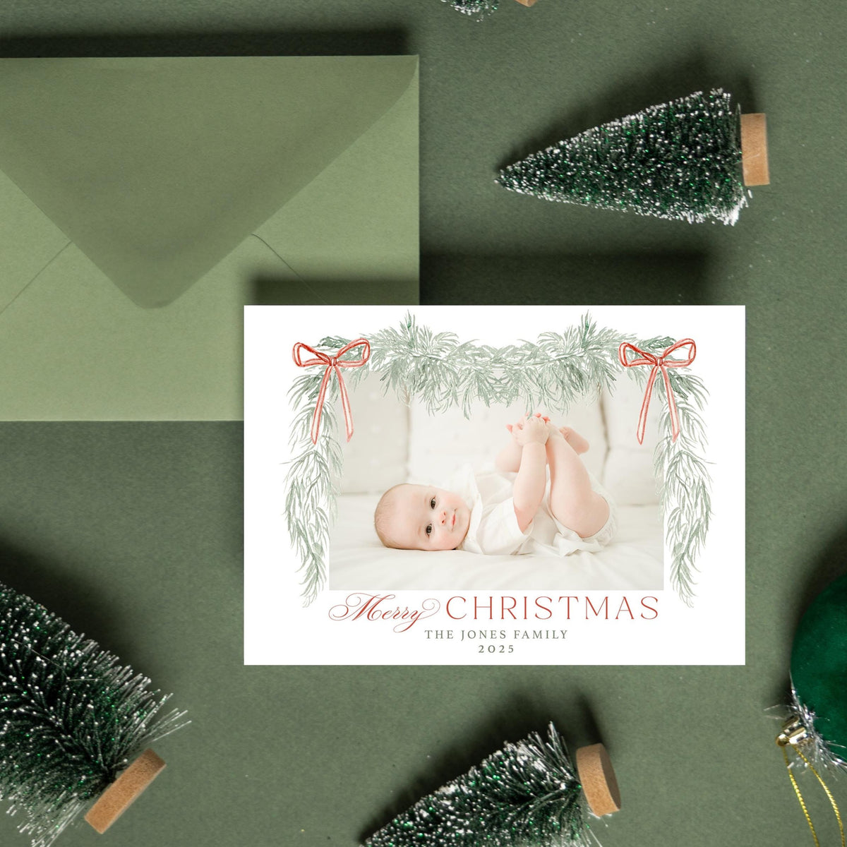 A green envelope, a Christmas card with a photo of a baby, and some small green Christmas trees are arranged on a green background.