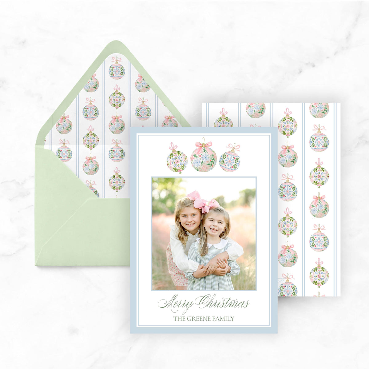 A Christmas card featuring two young girls smiling and embracing, with a pattern of Christmas ornaments on the envelope and card.
