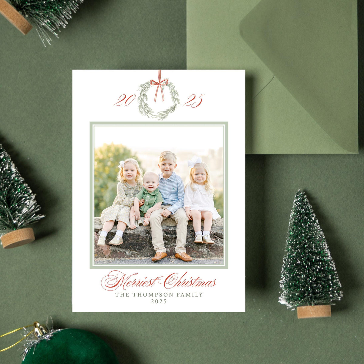 a green envelope with a photo of three children sitting on a rock, surrounded by Christmas decorations such as small trees and ornaments.