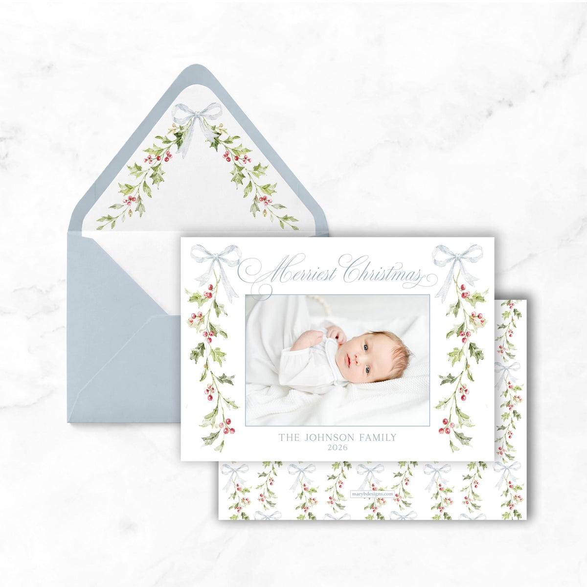 A Christmas card with a photo of a baby lying on a bed, surrounded by a floral design and a bow. The card is placed inside an envelope with a similar floral design.