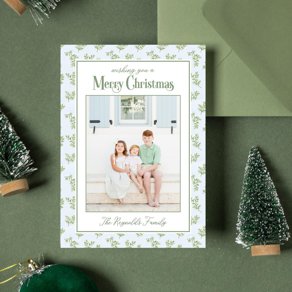 a Christmas card with a photo of a family, surrounded by Christmas decorations such as small trees, ornaments, and a green envelope.