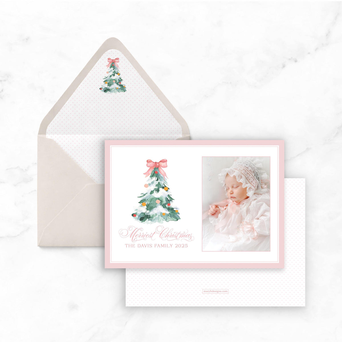 A pink and white Christmas card with a photo of a baby and a Christmas tree on it, placed on a marble surface.