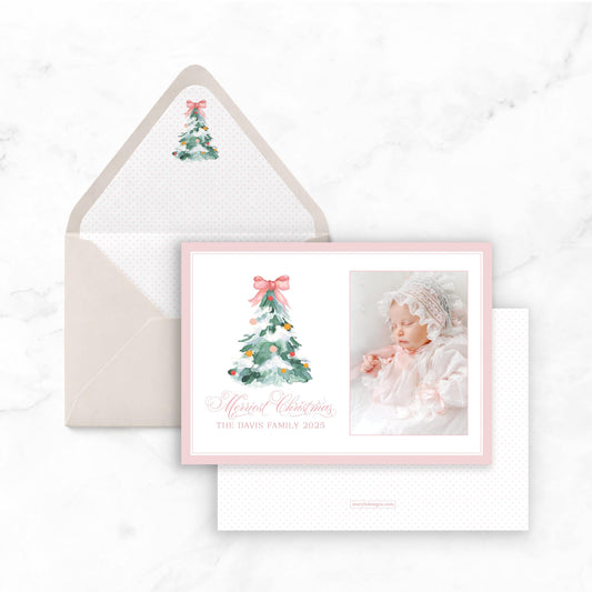 A pink and white Christmas card with a photo of a baby and a Christmas tree on it, placed on a marble surface.
