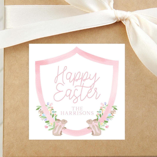 May include: A pink and white Easter gift tag with a pink shield design. The tag has the text 'Happy Easter' in pink script font and the text 'The Harrisons' in black font. The shield is surrounded by a floral design with two bunnies wearing pink bows.