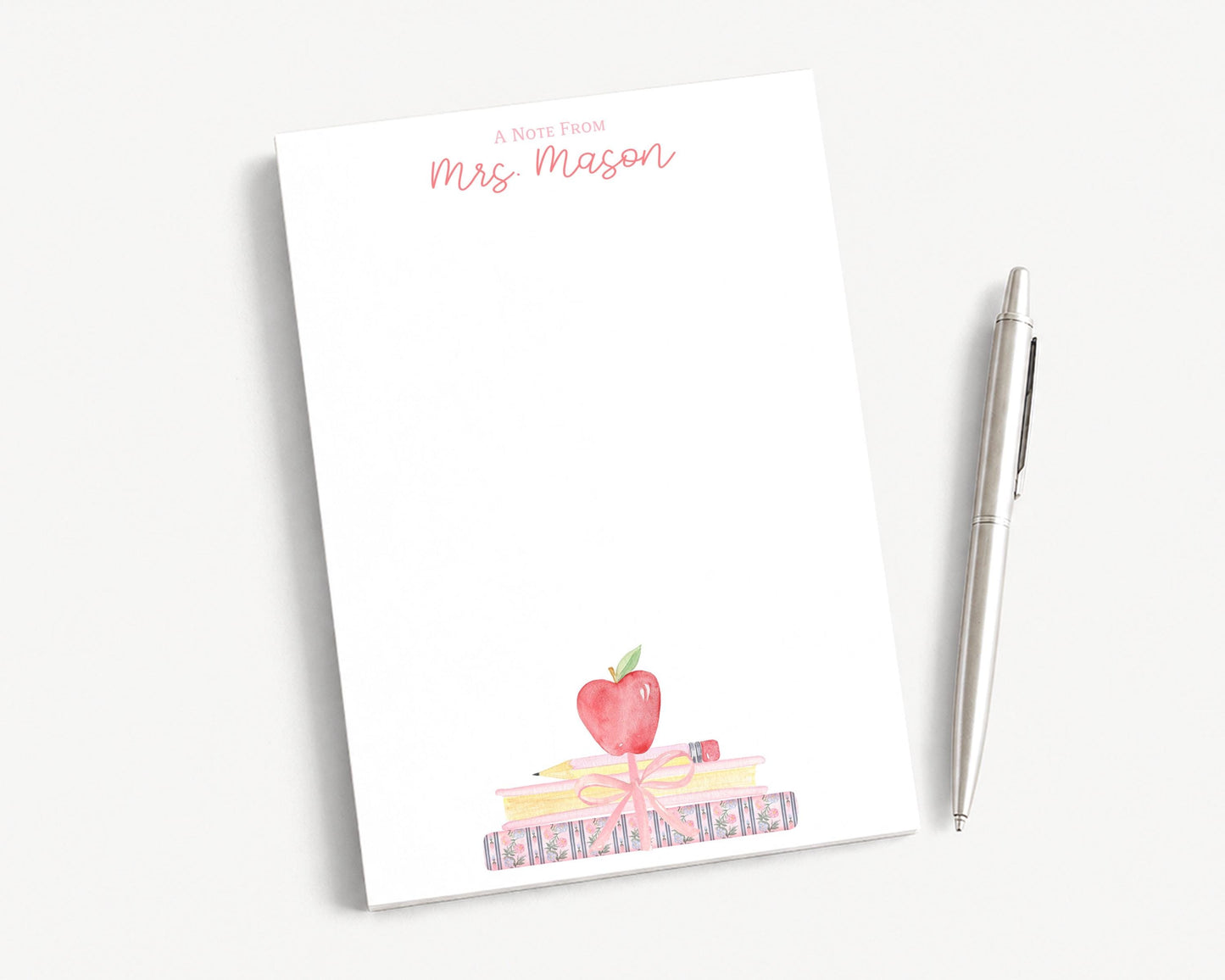 A blank white notepad with a watercolor illustration of a stack of books and an apple on top, accompanied by a pen on a white surface.