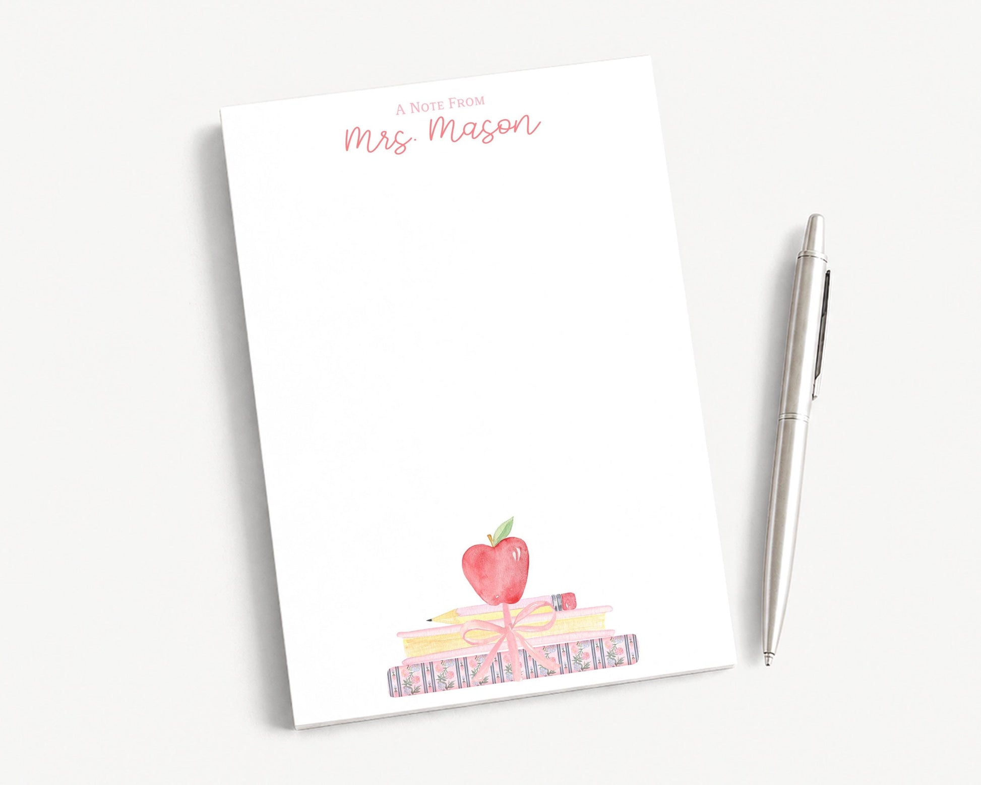A blank white notepad with a watercolor illustration of a stack of books and an apple on top, accompanied by a pen on a white surface.