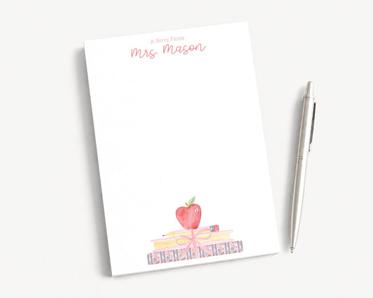 A blank white notepad with a watercolor illustration of a stack of books and an apple on top, accompanied by a pen on a white surface.