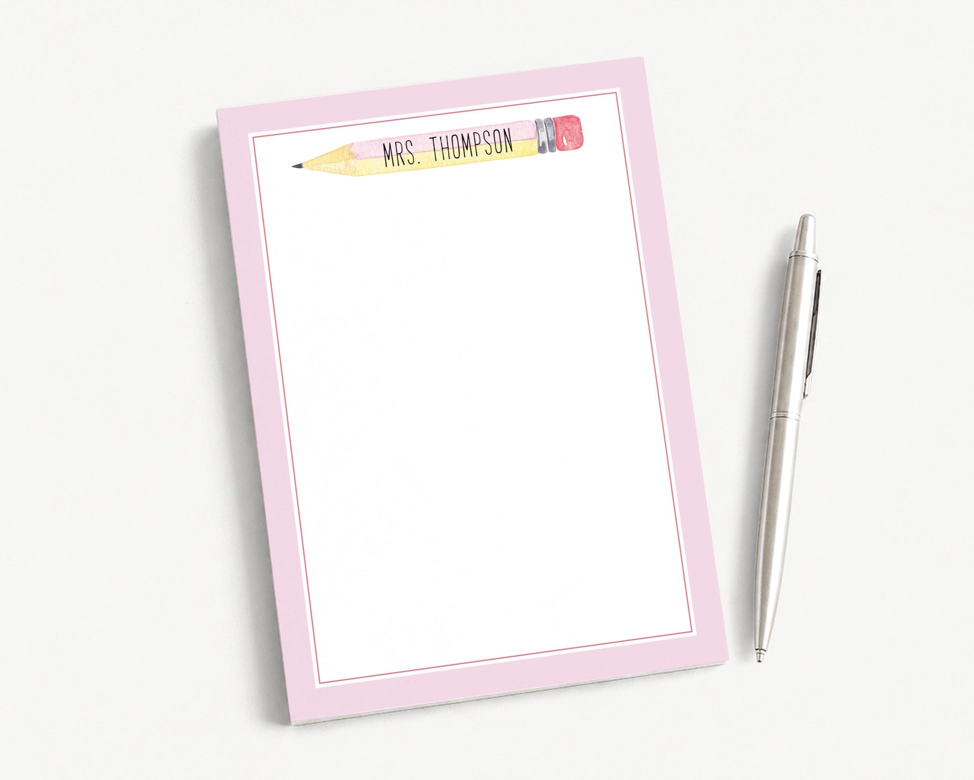 A pink-framed notepad with a pencil on top, accompanied by a pen, is placed on a white surface.